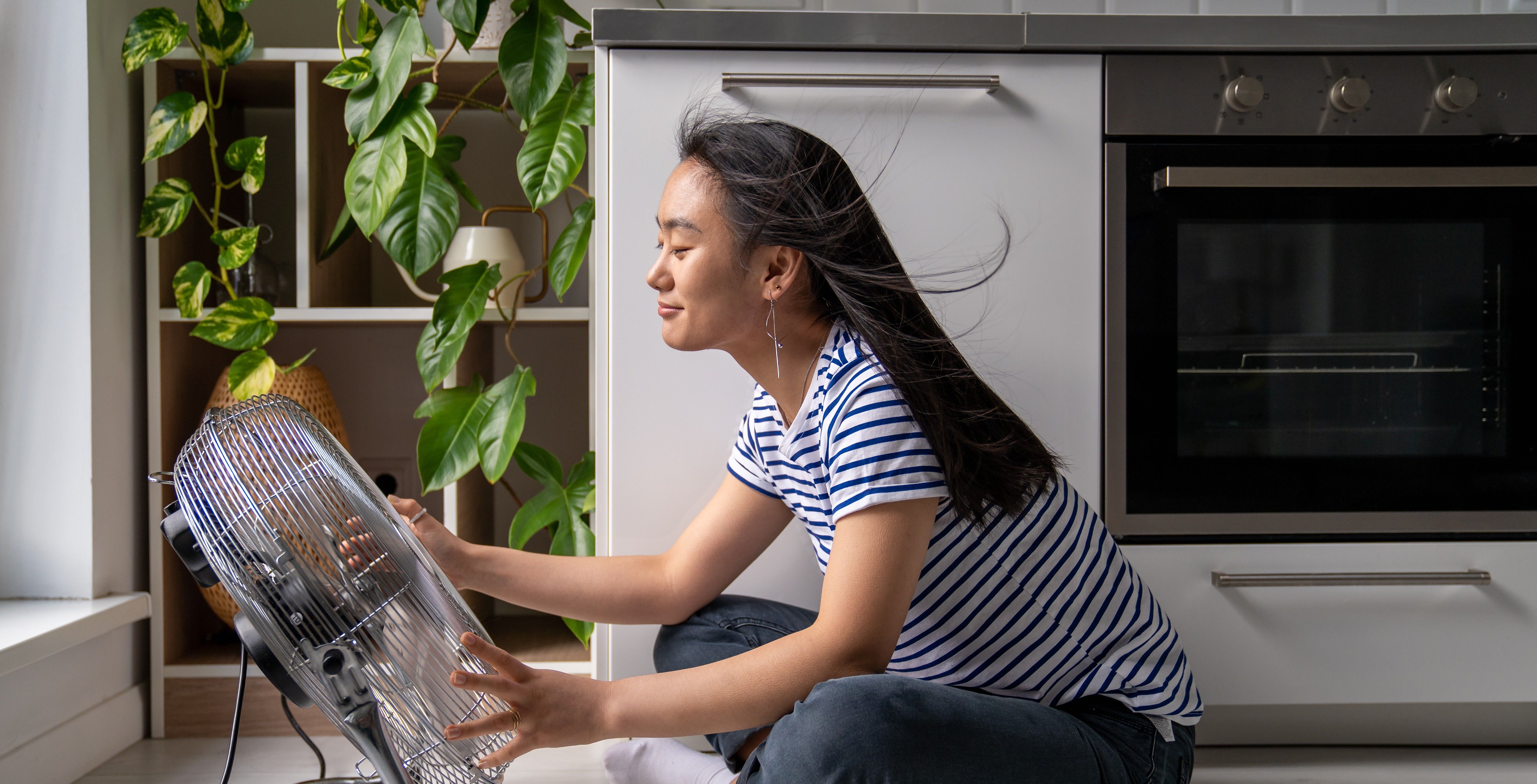Junge Frau sitzt vor Ventilator an heißem Sommertag, um die Wohnung ohne Klimaanlage abzukühlen.