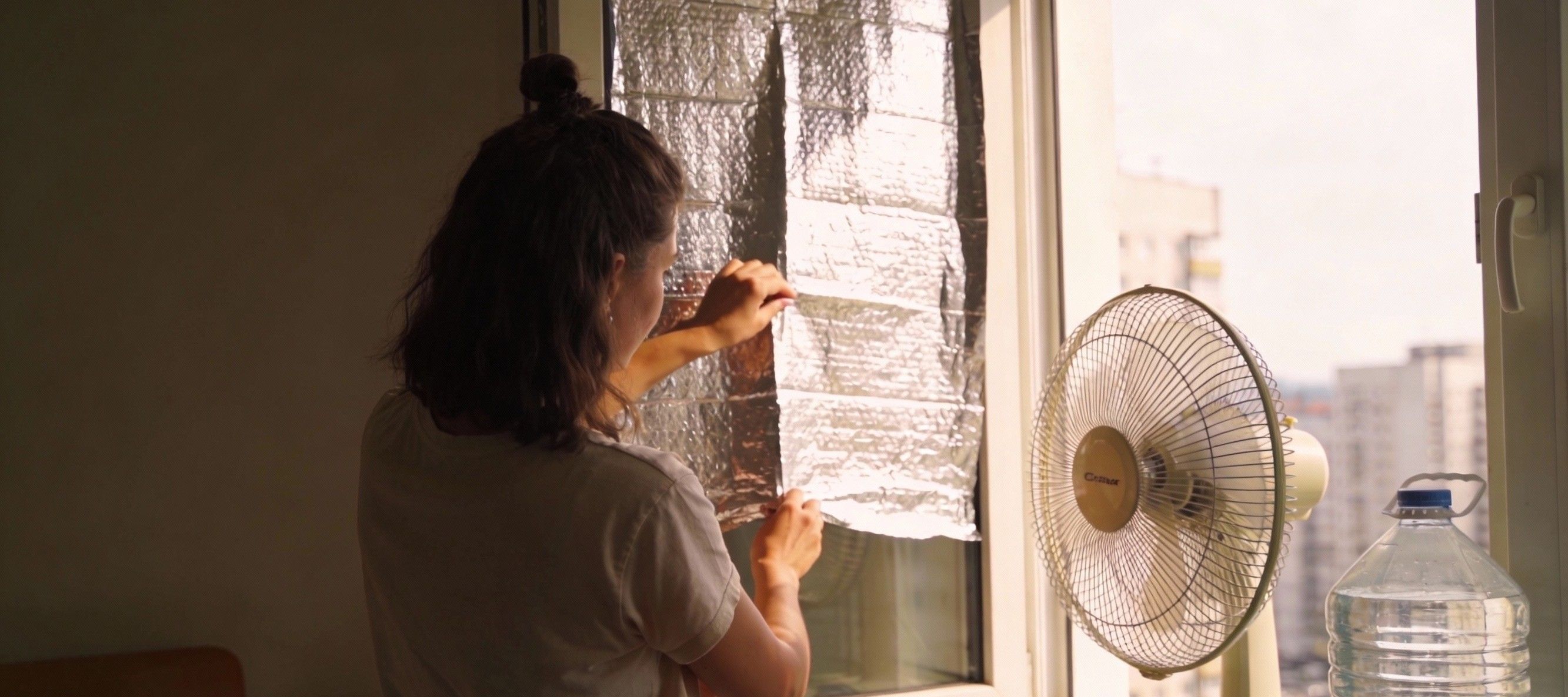 Junge Frau dämmt im Sommer ihr Fenster mit Abdeckfolie, um die Wohnung abzukühlen.