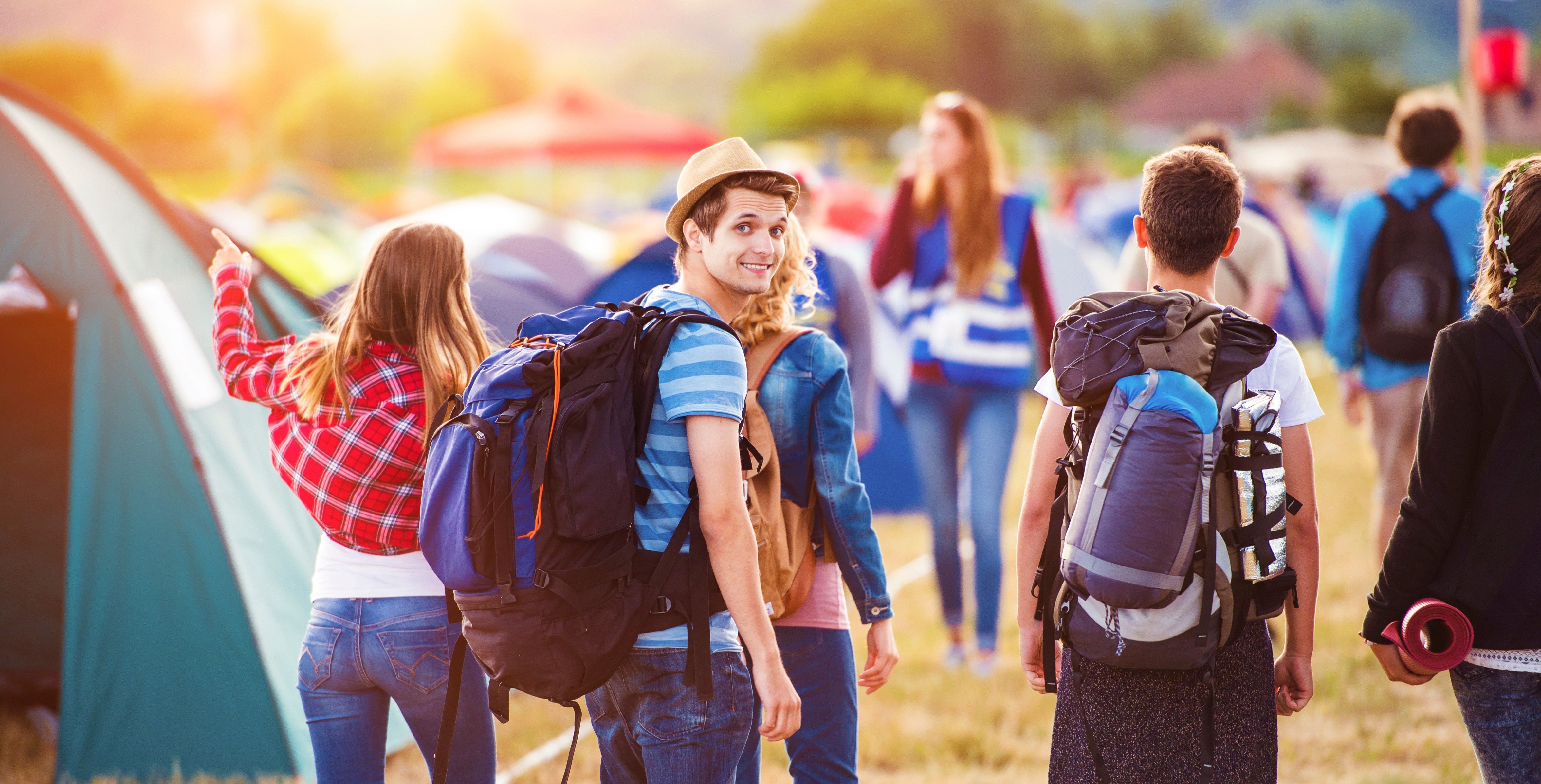 Junge Freundesgruppe auf einem sommerlichen Zeltplatz eines Festivals mit gepackten Rucksäcken.