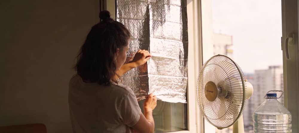Junge Frau dämmt im Sommer ihr Fenster mit Abdeckfolie, um die Wohnung abzukühlen.