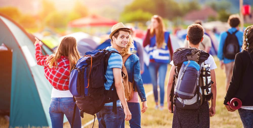 Junge Freundesgruppe auf einem sommerlichen Zeltplatz eines Festivals mit gepackten Rucksäcken.