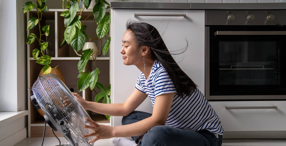 Junge Frau sitzt vor Ventilator an heißem Sommertag, um die Wohnung ohne Klimaanlage abzukühlen.