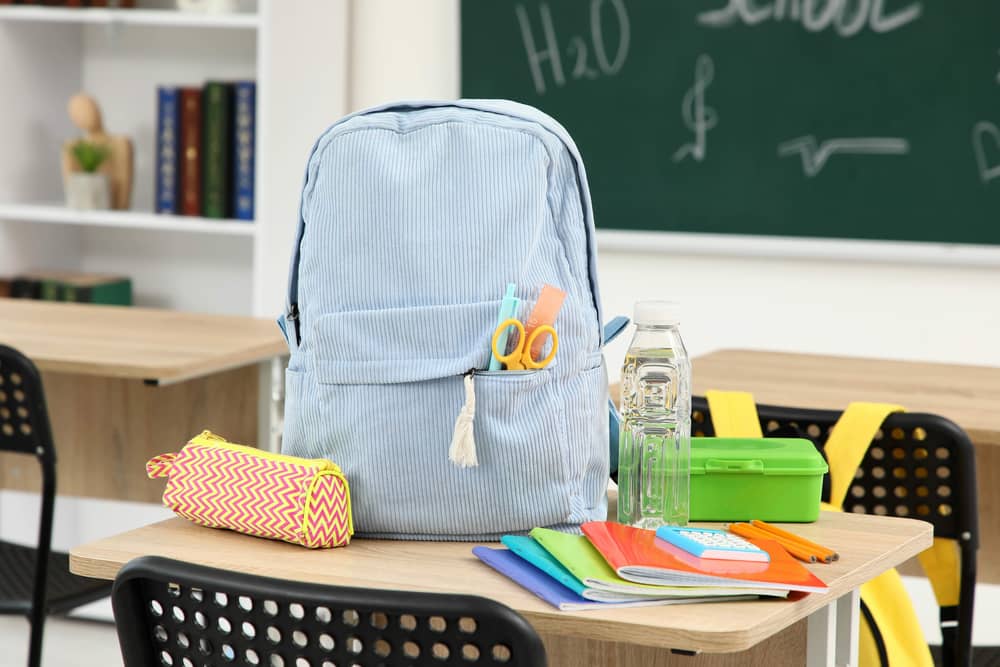 Ein Schulrucksack auf einem Tisch in einem Klassenraum, daneben liegen Federtasche, Hefte, eine Brotdose und eine Wasserflasche.