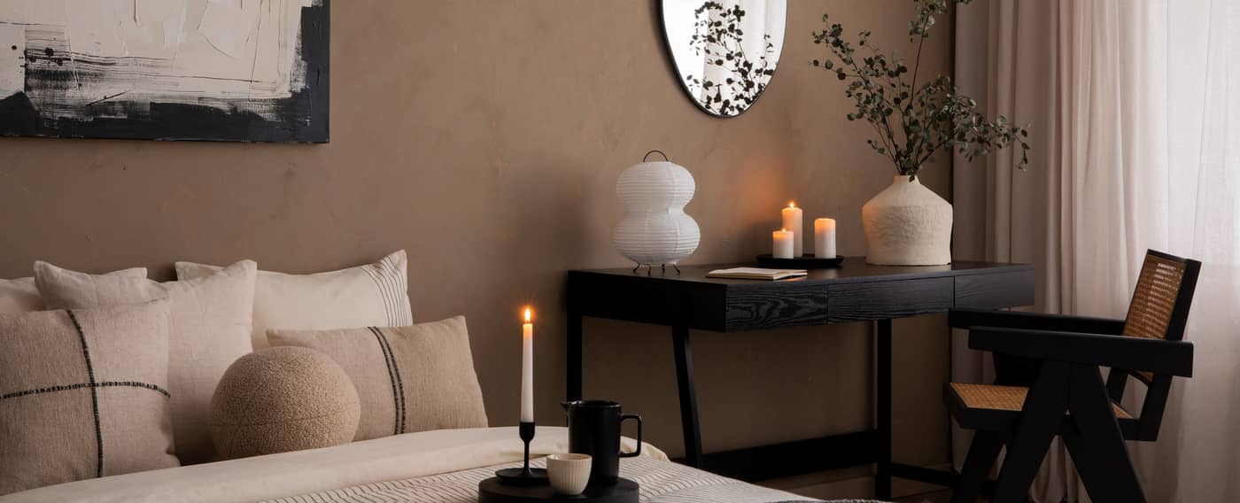 In einem Schlafzimmer stehen ein Bett, ein Schminktisch und ein Stuhl. Der Raum sowie die Möbel und Dekoelemente sind in Erdtönen gehalten.