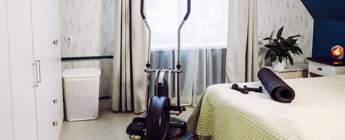 In einem Schlafzimmer sind mehrere Sportgeräte: Ein Crosstrainer steht au dem Boden. Auf dem Bett liegen kleine Hanteln sowie eine zusammengerollte Yogamatte