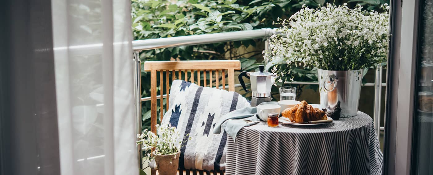 Balkon mit kleinem Tisch, Sitzkissen, Pflanzen und gedecktem Frühstück, gesehen durch geöffnete Vorhänge.