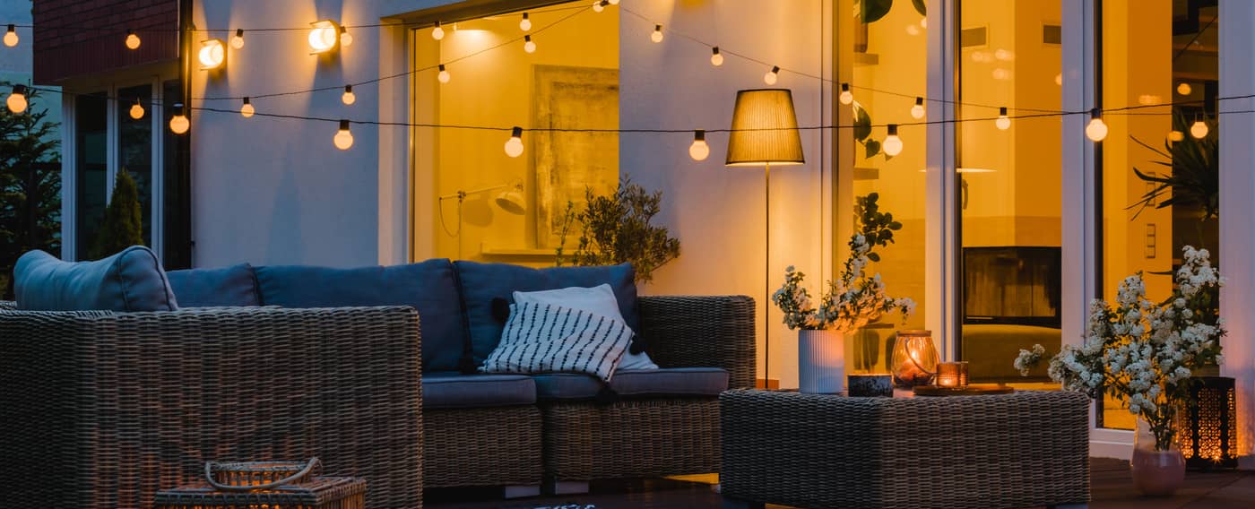 Eine schöne Terrasse vor einem Einfamilienhaus im Abendlicht. Über Gartenmöbeln hängen leuchtende Lichterketten