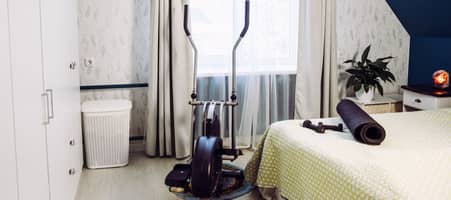 In einem Schlafzimmer sind mehrere Sportgeräte: Ein Crosstrainer steht au dem Boden. Auf dem Bett liegen kleine Hanteln sowie eine zusammengerollte Yogamatte