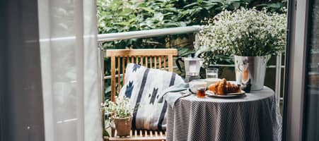 Balkon mit kleinem Tisch, Sitzkissen, Pflanzen und gedecktem Frühstück, gesehen durch geöffnete Vorhänge.