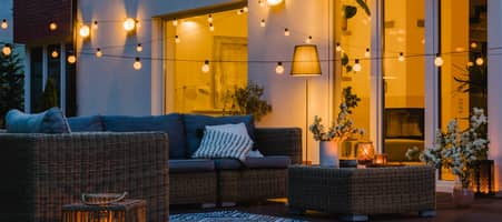 Eine schöne Terrasse vor einem Einfamilienhaus im Abendlicht. Über Gartenmöbeln hängen leuchtende Lichterketten