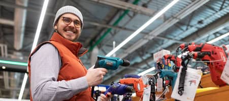 Ein Mann steht mit breitem Lächeln in einem Baumarkt und hält einen Schraubenzieher